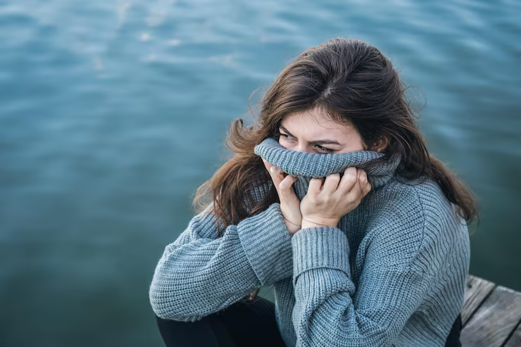 A woman in a grey jumper sits by water, pulling her collar over her face