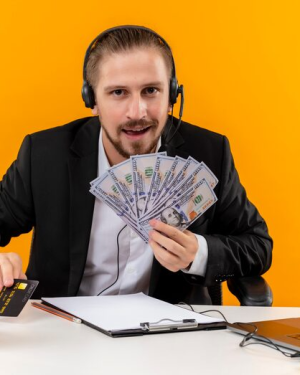 Man in suit with headset holding cash and credit card at desk with laptop and stationery against orange background
