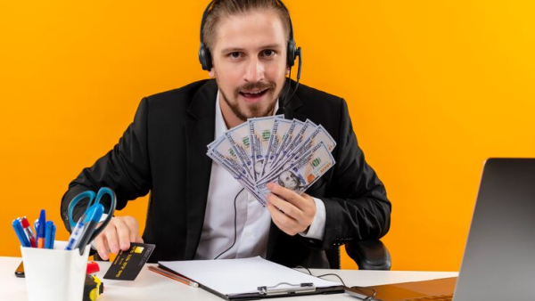 Man in suit with headset holding cash and credit card at desk with laptop and stationery against orange background