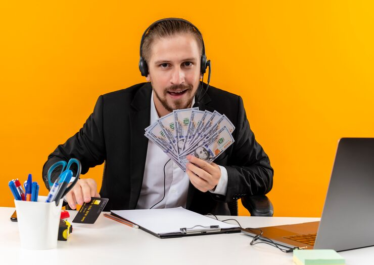 Man in suit with headset holding cash and credit card at desk with laptop and stationery against orange background