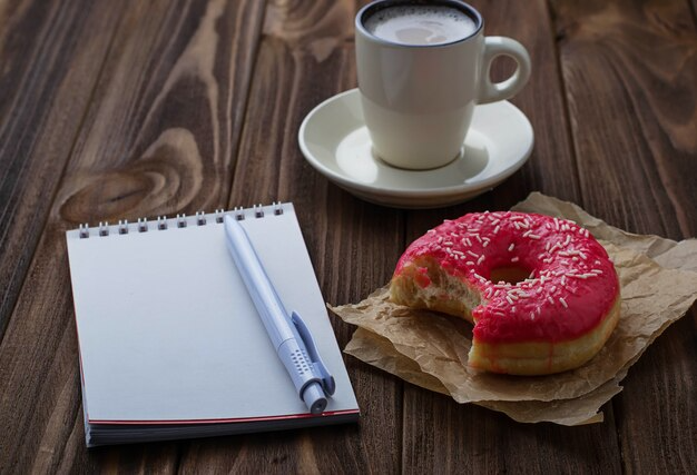 Open notebook with pen next to bitten pink doughnut and cup of hot drink on wooden table